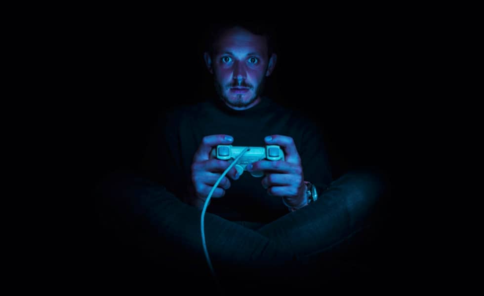 A young man holding a video game controller, lit only by the blue hue of a video game screen