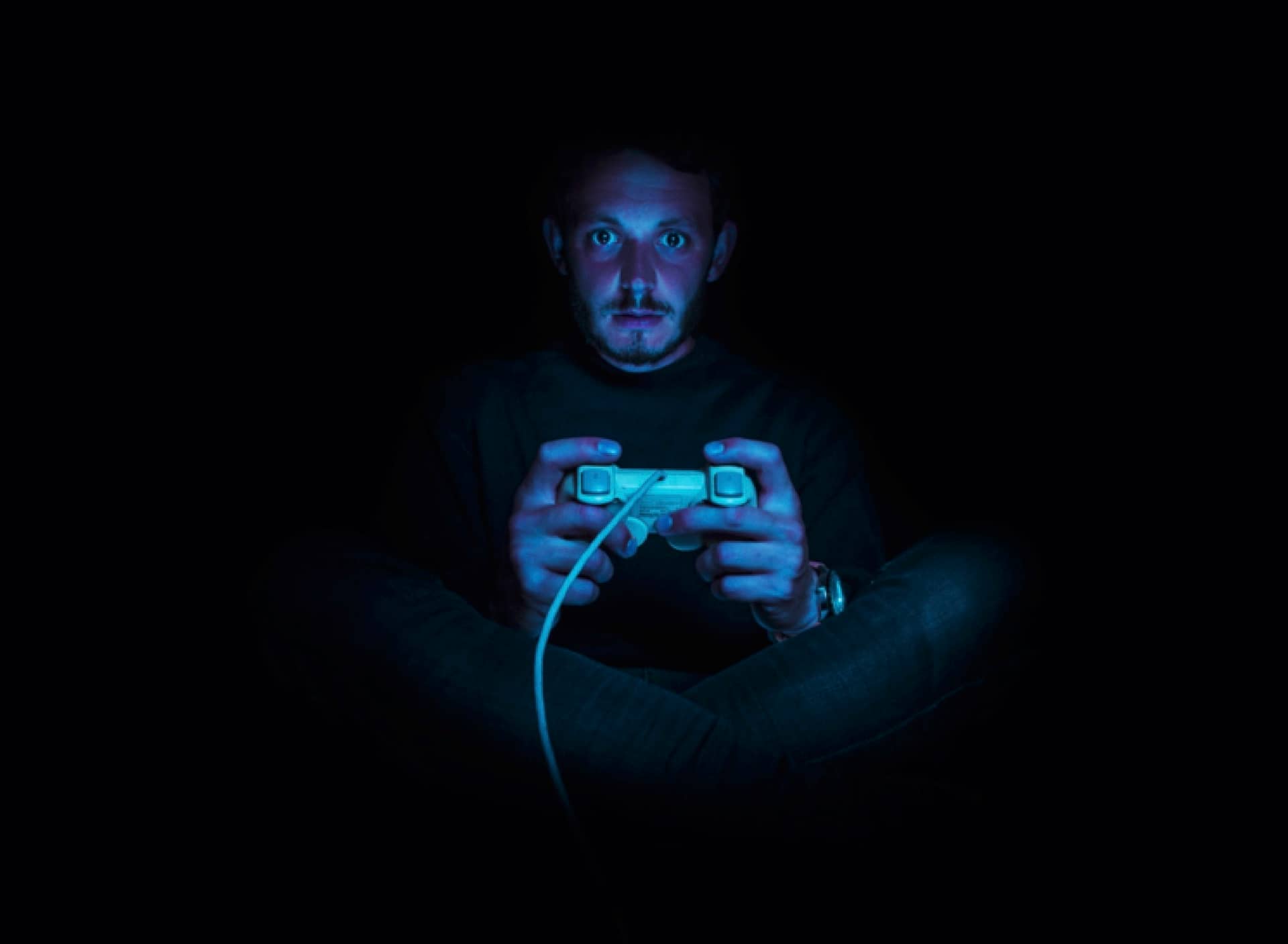 A young man holding a video game controller, lit only by the blue hue of a video game screen